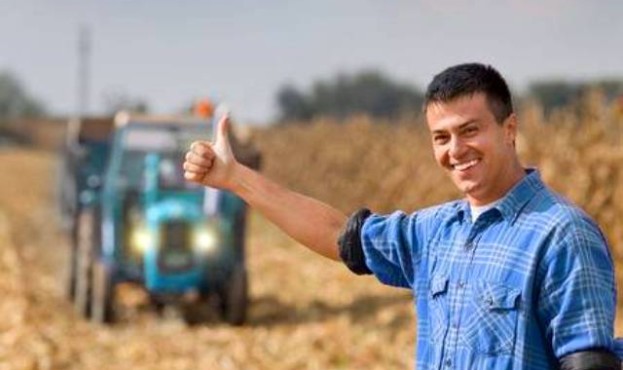49205422-junger-lächelnder-landwirt-mit-daumen-nach-oben-als-ok-zeichen-auf-ackerland-traktor-mit-anhänger-im-hin
